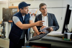 mid-adult-manager-car-mechanic-talking-while-using-computer-auto-repair-shop-scaled